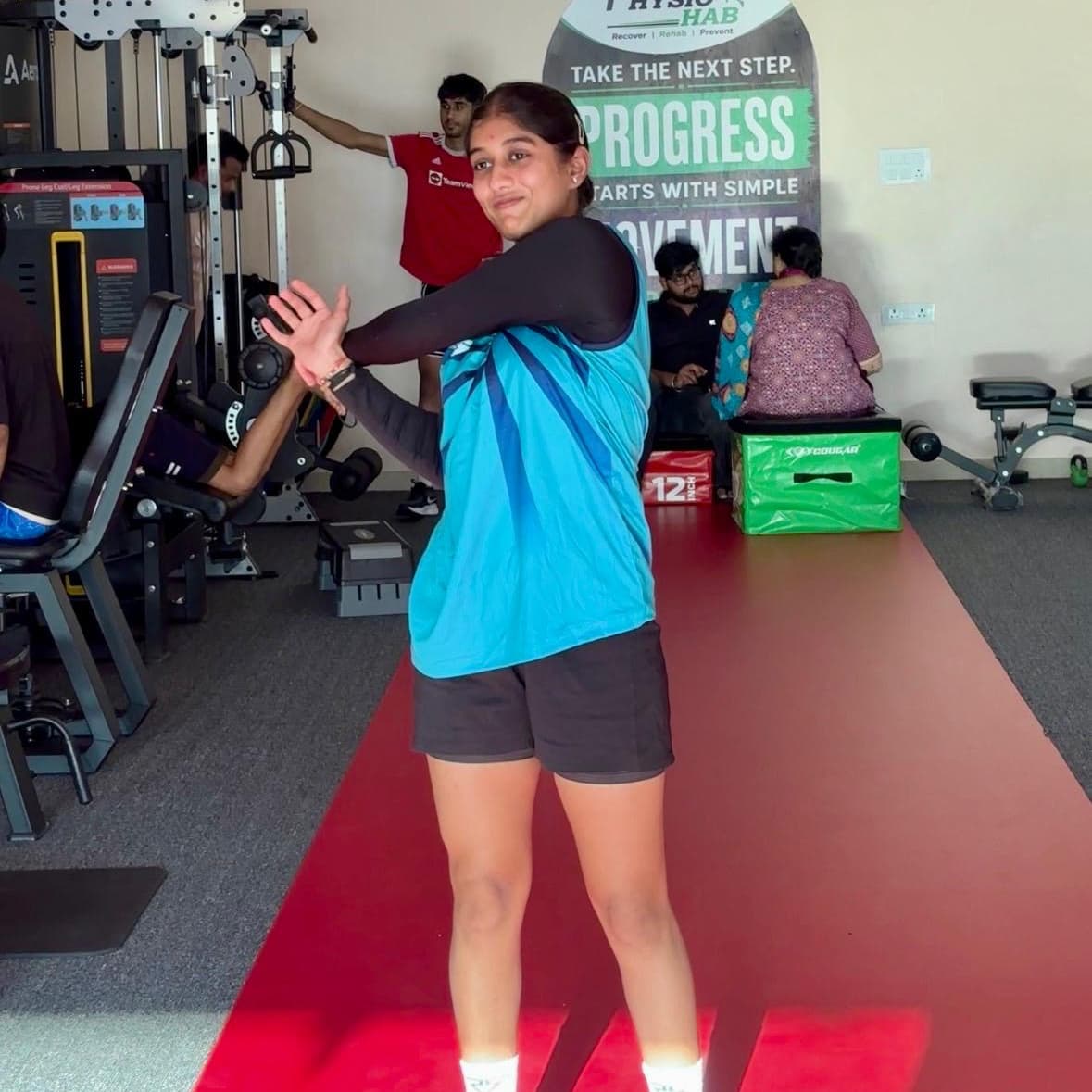 Patient performing a standing shoulder and arm stretch on the clinic floor between exercise stations