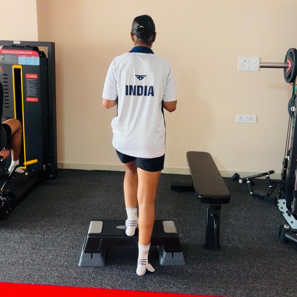 Patient performing a single-leg step-up for balance and strength training near the hip abduction-adduction machine