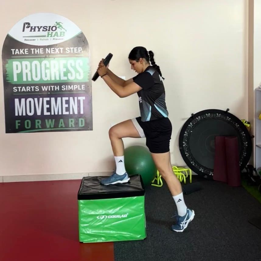 Patient performing a weighted step-up onto a plyo box beside the clinic's 'Progress starts with simple movement' wall graphic